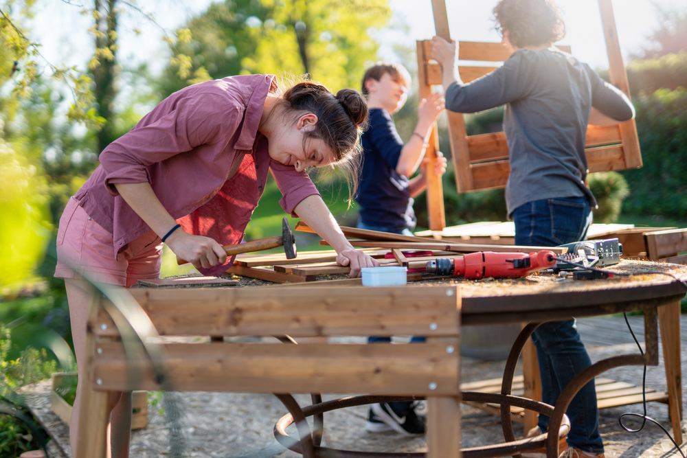 Two teenagers helping their parent build a wooden garden planter on a sunny day.
